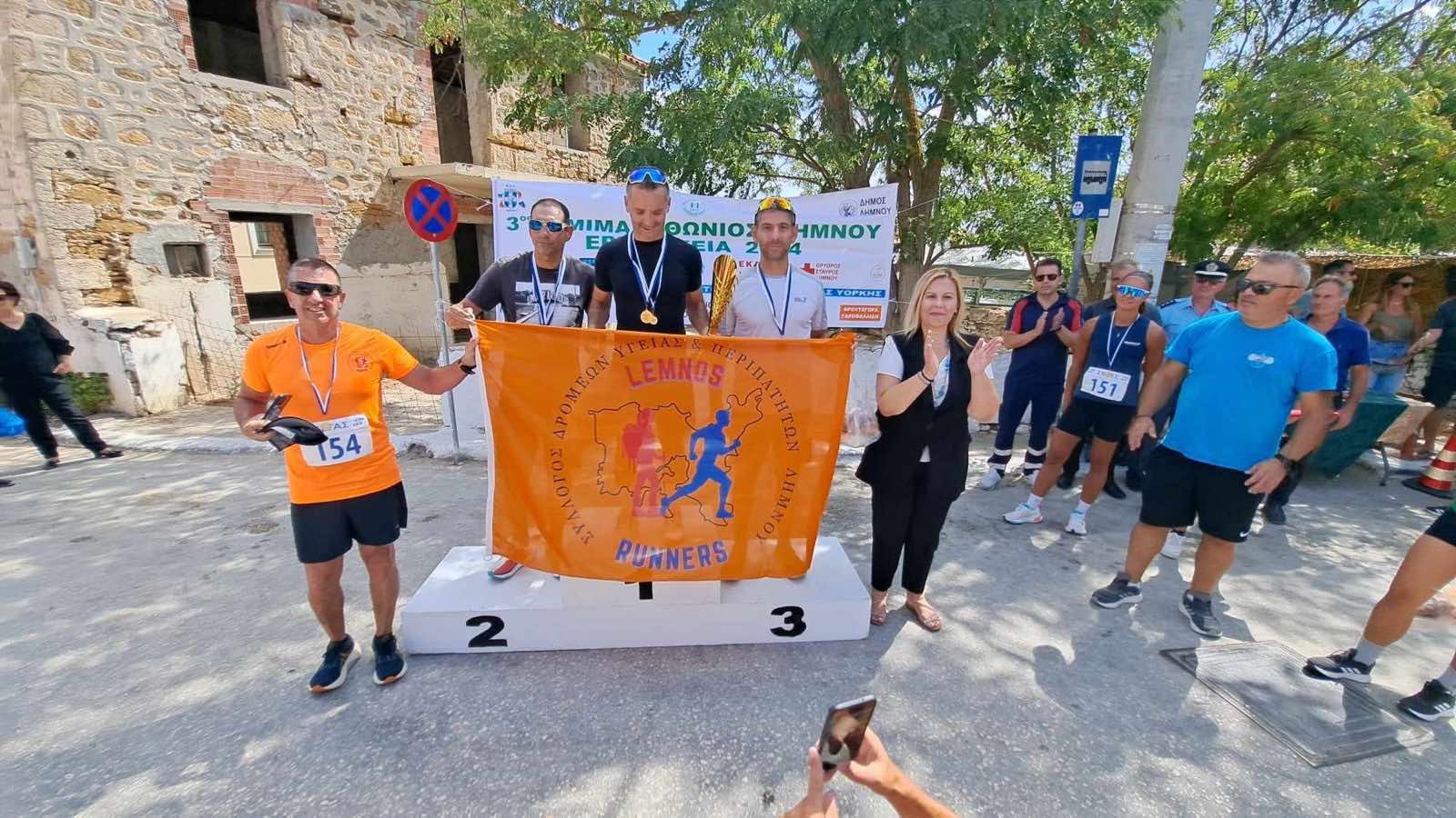 Απονομή στο βάθρο στην πλατεία Κοντοπουλίου, με νικητές, μετάλλια, πορτοκαλί σημαία και επισήμους.